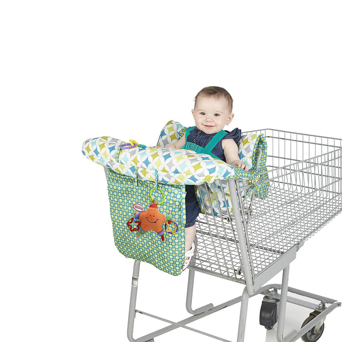 A smiling baby sits in a supermarket cart, enjoying the Nuby 2-in-1 Universal Size Shopping Cart & High Chair Cover with patterned fabric and a hanging orange toy, adding comfort and fun against a crisp white background.
