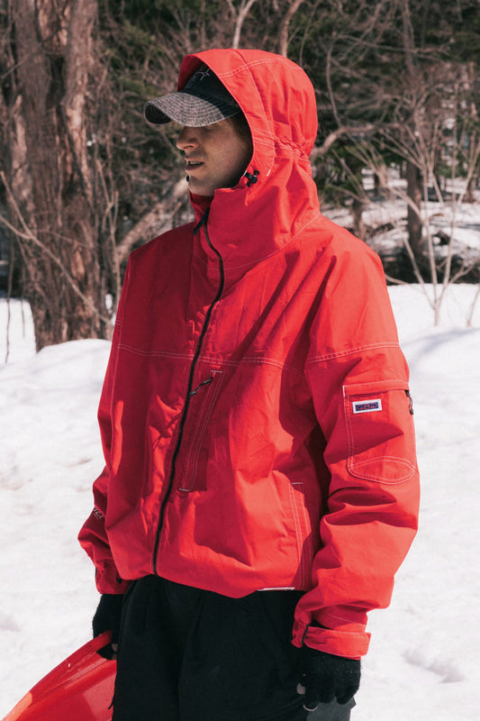 A person wearing a 686 Dojo® GORE-TEX Jacket, black gloves, and a gray cap stands outdoors in the snow holding a red object, with trees in the background.