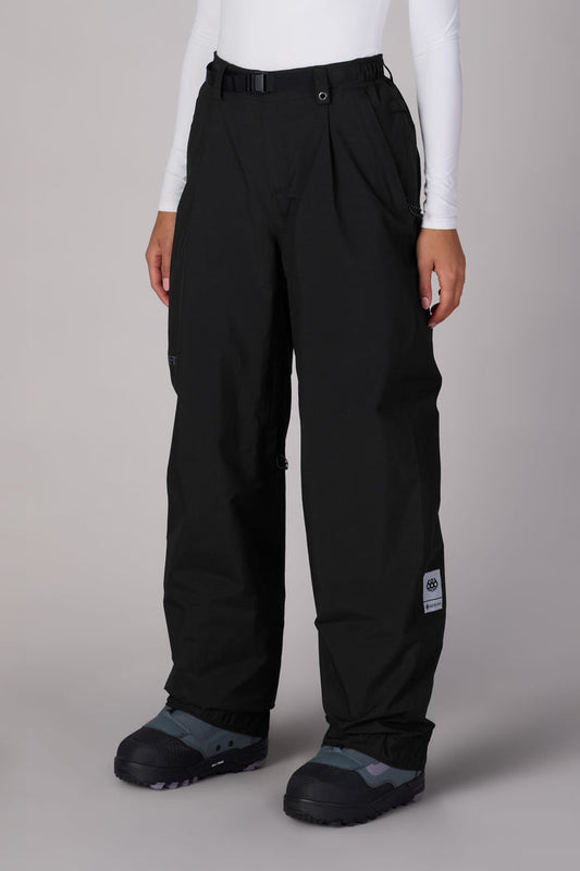 A person wears 686 Women's GORE-TEX Outline™ Shell Pant by 686—loose-fitting black waterproof pants with a belt and ankle patch—paired with a white long-sleeve top and black snow boots, standing on a neutral background.