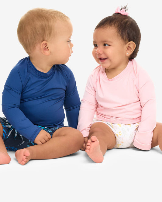 Two babies, one in a blue long-sleeve crew neck top and the other in a pink one, sitting and smiling.
