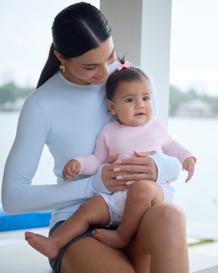 Woman in a blue long-sleeve top carrying a baby with a pink long-sleeve top outside by the ocean.