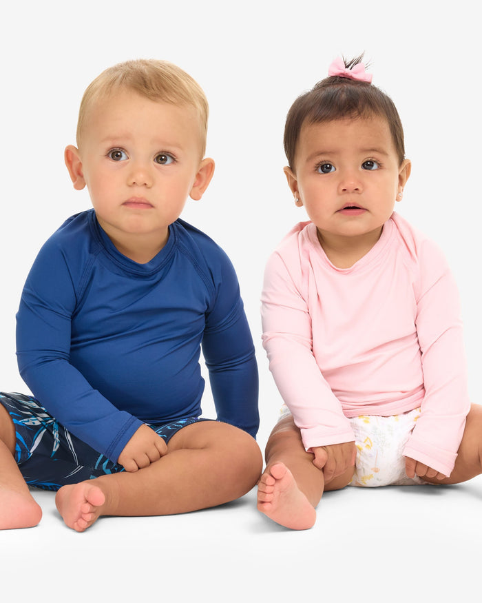 Two babies, one in a blue long-sleeve crew neck top and the other in a pink one, sitting and smiling.