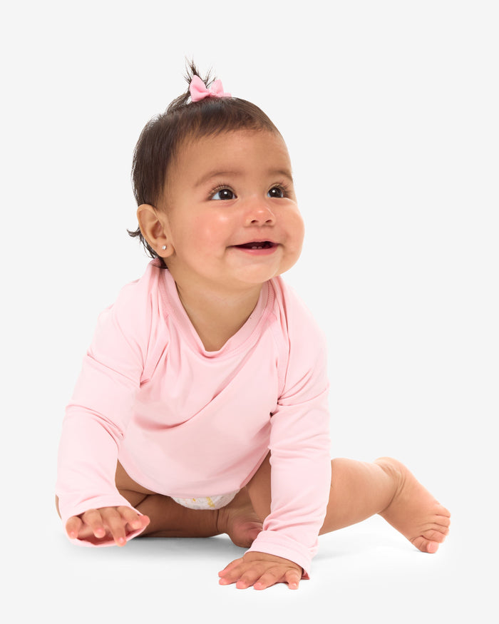 A baby in a pink long-sleeve sitting and smiling in front of a white background.