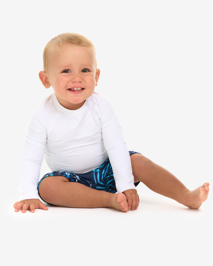 A baby in a white long-sleeve sitting and smiling in front of a white background.
