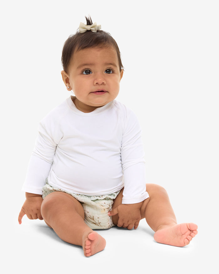 A baby in a white long-sleeve sitting and smiling in front of a white background.