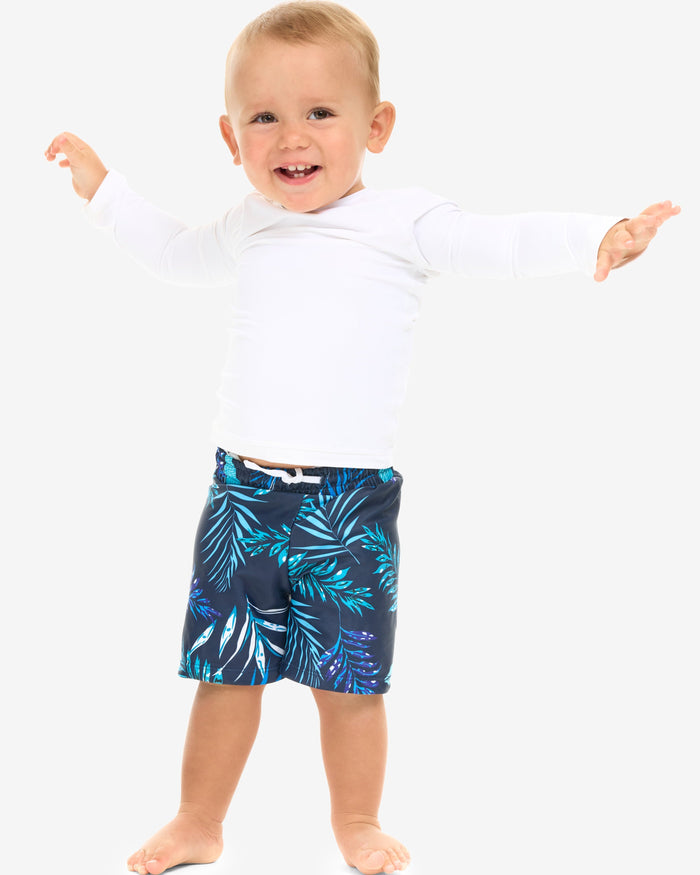 A baby in a white long-sleeve standing and smiling in front of a white background.