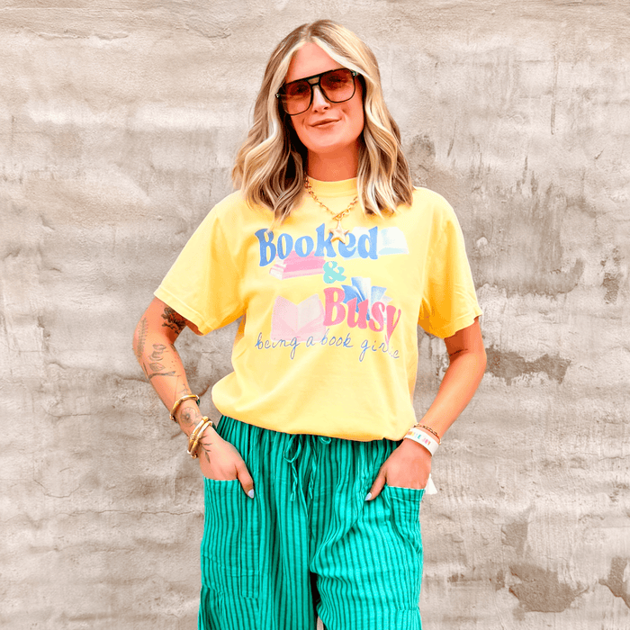 Yellow Booked & Busy tee with colorful text, worn by a woman with sunglasses and green striped pants against a textured wall.