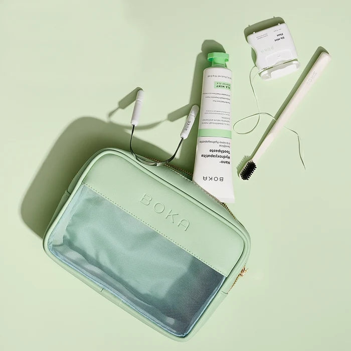 Mint green travel case with Boka branding, including a tube of cream, hairbrush, and other items on a light green background.