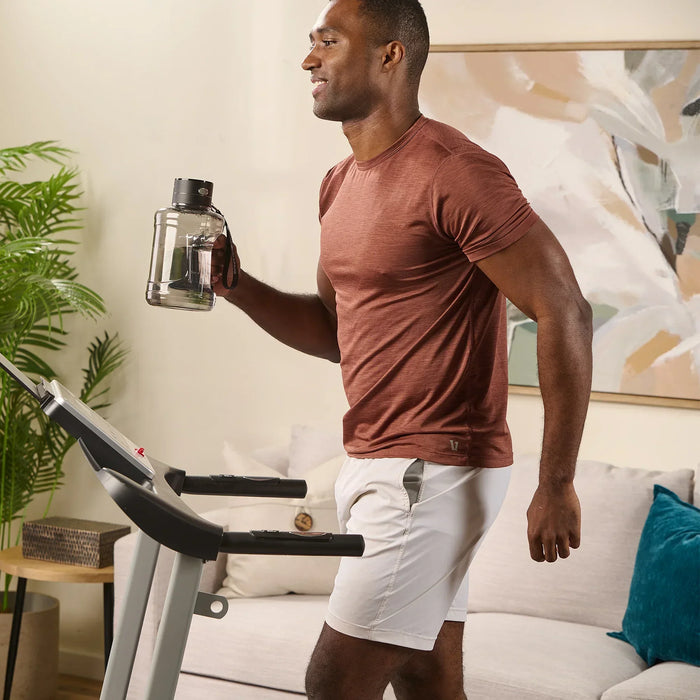 Man walking on a treadmill in a rust-colored shirt, holding the HydroLife Chug bottle by its wrist strap.