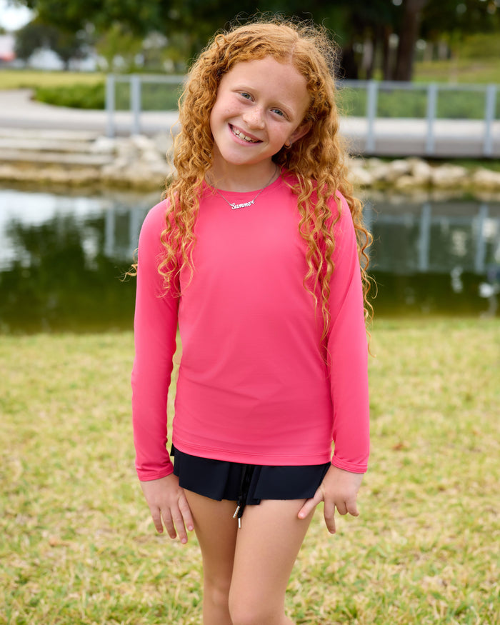 Young girl in a pink long-sleeve shirt and black shorts standing outdoors by a body of water.