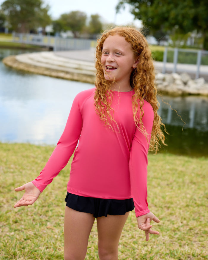 Girl in pink long-sleeve shirt and black shorts standing outdoors by a lake.