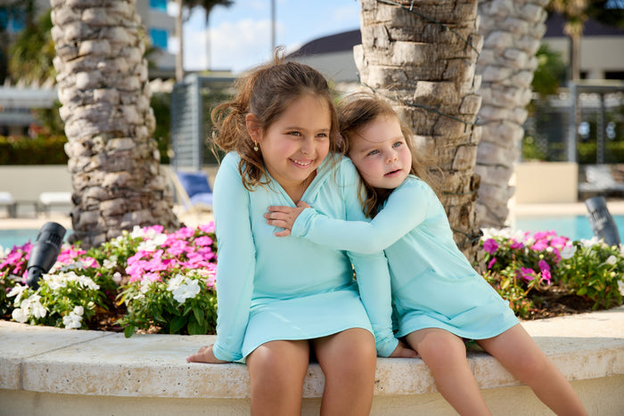 Two young girls sitting on a bench, wearing matching long-sleeve hoodie dresses in a light color, with flowers and palm trees in the background.