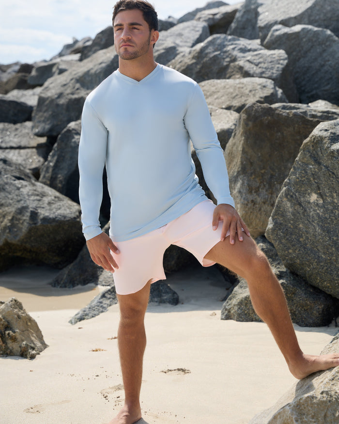 Man in light blue long-sleeve shirt and pink shorts standing on a rocky beach.