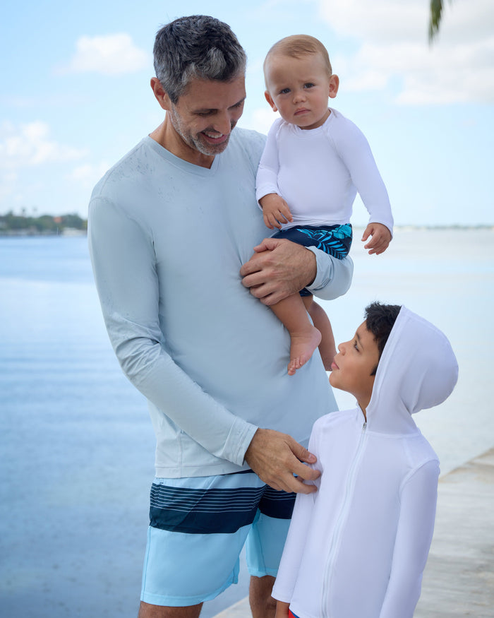 A man wearing a gray long-sleeved V-neck shirt, with two kids, by the ocean.