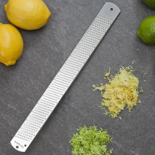 A Microplane grater/zester lying on a surface with lemon and lime zest around it, and a lime and lemon partially visible at the top left corner.