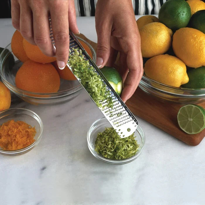 Person zesting a lime over a bowl with lemons and oranges on a marble surface.