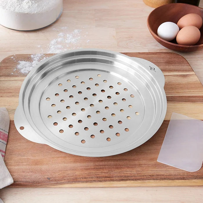 A stainless steel spaetzle top, featuring a perforated grater-style lid and a flanged design for easy grip, accompanied by a dough scraper on a wooden table with flour and eggs.