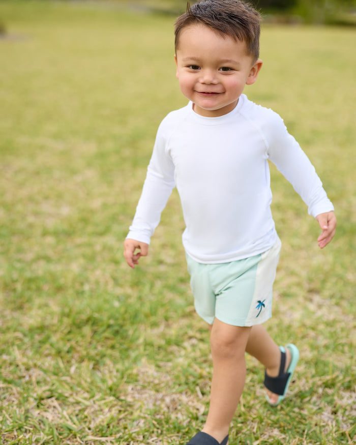 Child wearing a white long-sleeve shirt and light green shorts standing on grass.