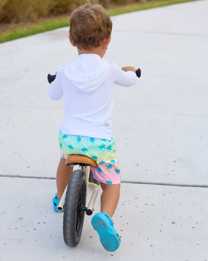 Child riding a balance bike on a sidewalk with grass and a fence in the background