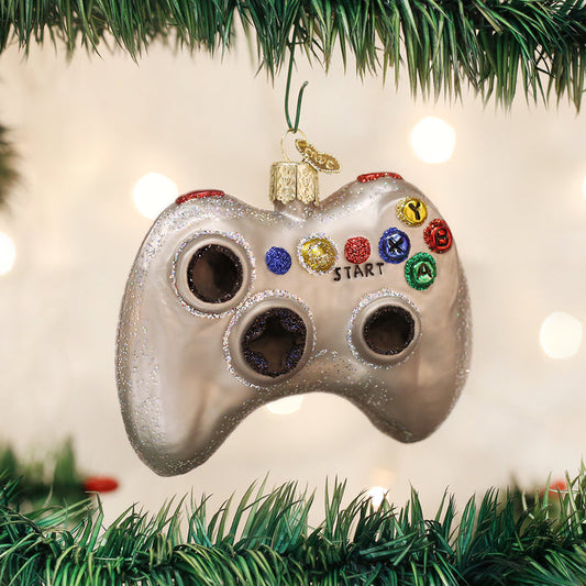 A Christmas tree ornament shaped like a video game controller hangs among green branches, with festive lights softly blurred in the background.