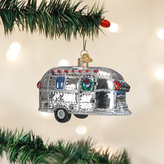 A shiny silver camper trailer ornament hangs on a green Christmas tree branch with festive lights softly glowing in the blurred background.