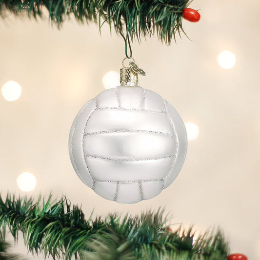 A white Christmas ornament designed to look like a volleyball hangs from a green pine tree branch, with soft white lights glowing in the blurred background.